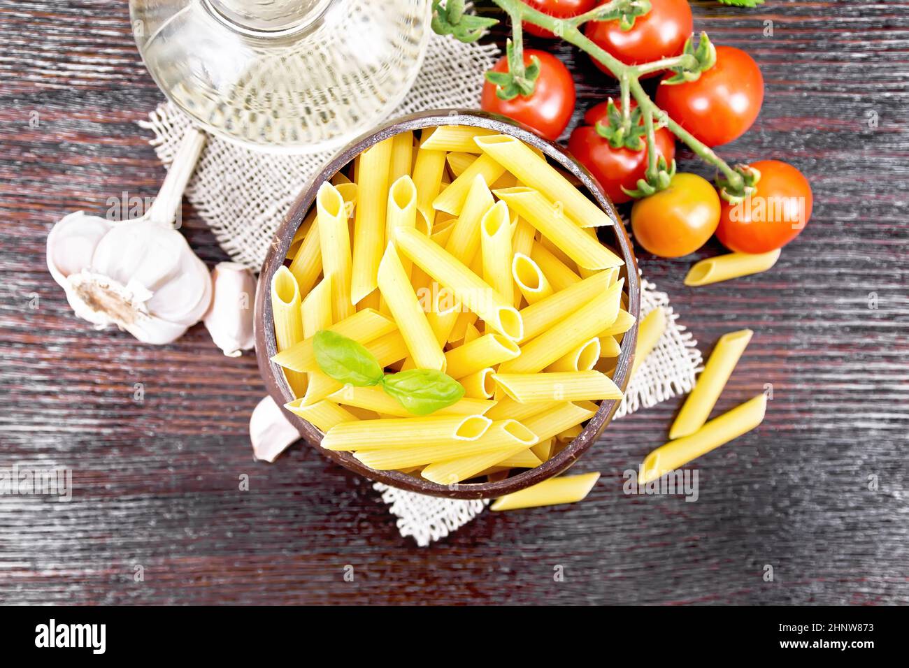 Wheat flour penne pasta in a bowl of coconut shells on sacking, tomatoes, garlic, vegetable oil in a decanter and parsley on wooden board background f Stock Photo