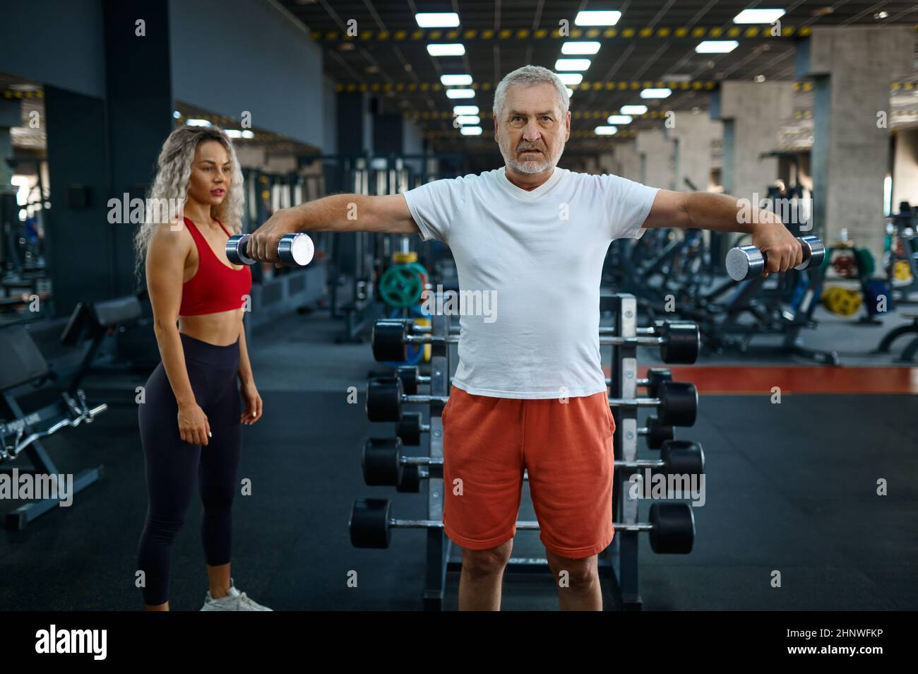 Old man doing exercise with dumbbells, female personal trainer, gym interior on background. Sportive grandpa with woman instructor, training in sport Stock Photo