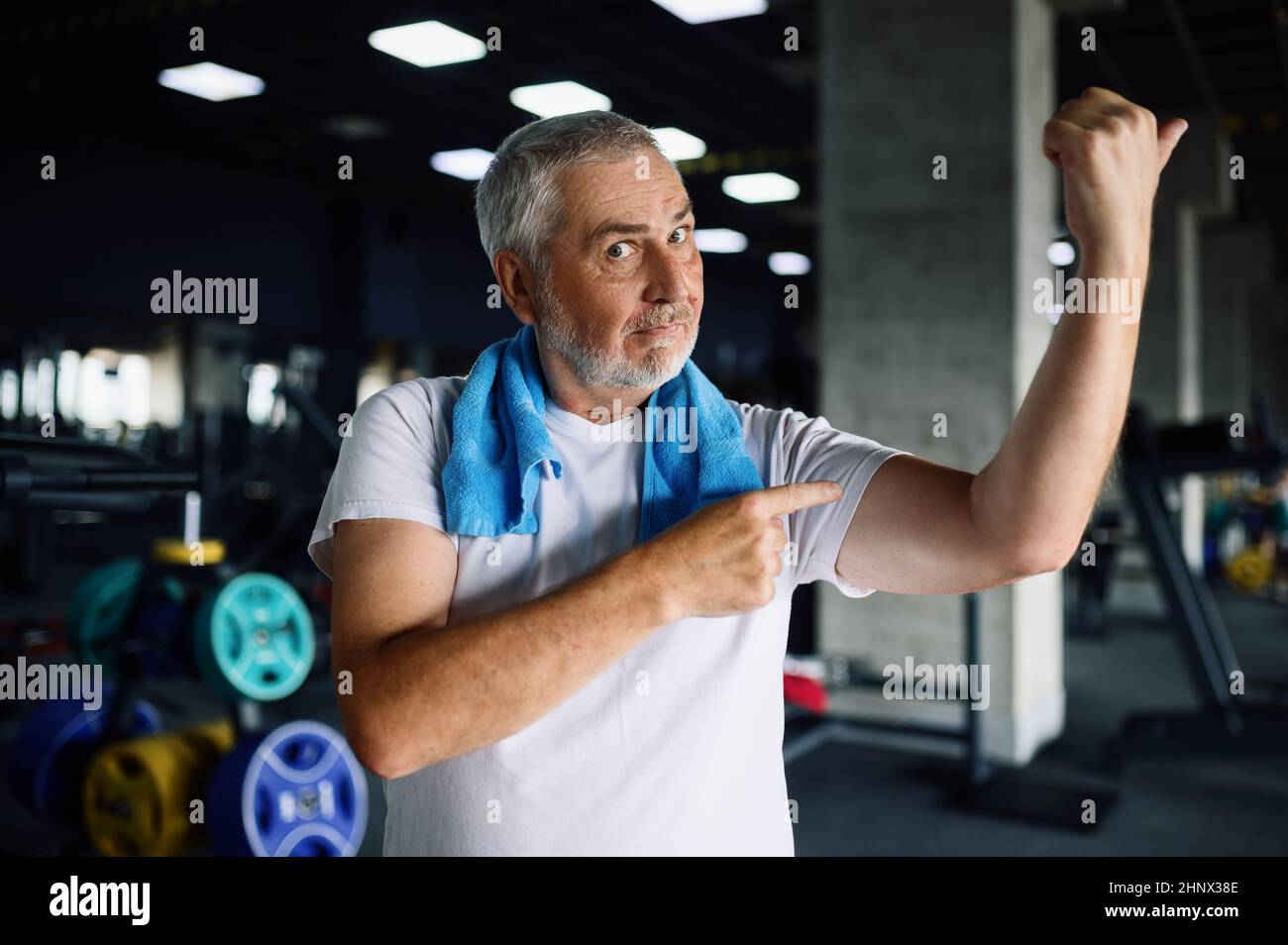 Happy old man shows his muscles, gym interior on background. Sportive