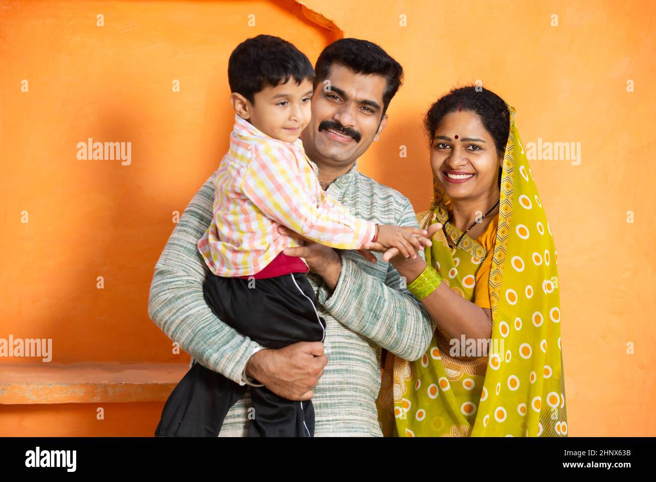 Happy young rural indian family laughing. father holding little child boy and mother wearing sari smiling,Couple wearing traditional cloths with kid. Stock Photo