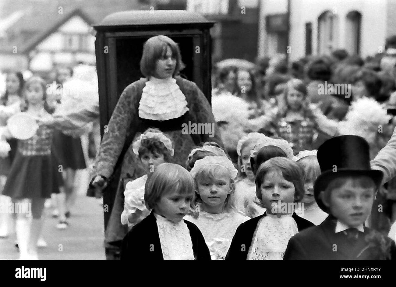 The annual Knutsford Royal May Day procession in 1976 in Knutsford, Cheshire, England, United Kingdom. It traditionally includes a fancy-dress pageant of children in historical or legendary costumes with  horse-drawn carriages. Here young children in historical costumes walk in the procession ahead of a teenage boy carrying the front handles of a litter or sedan chair. Stock Photo