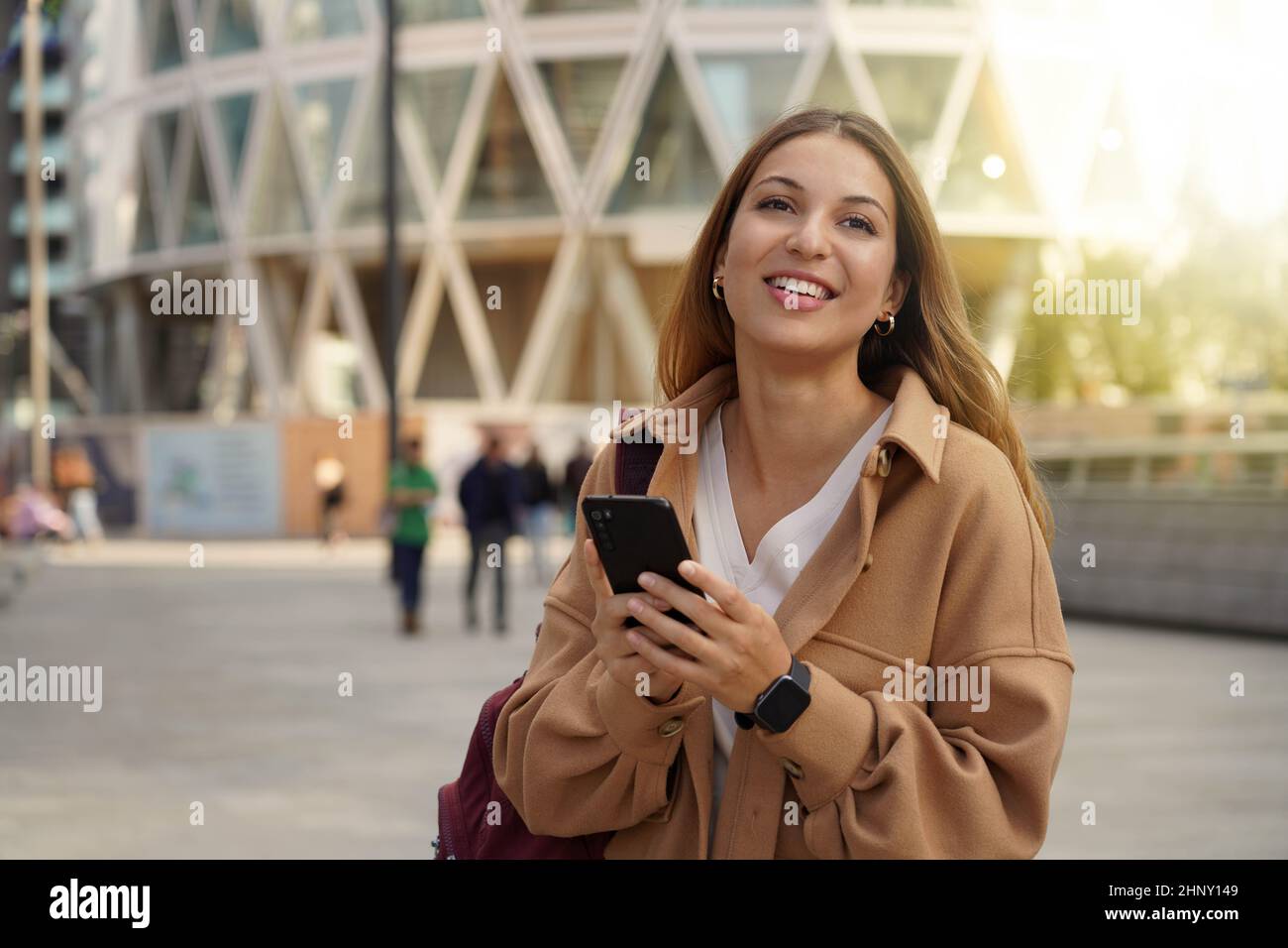 Casual cheerful girl walking and holding smart phone in city street wearing a coat in spring time. City lifestyle people technology. Stock Photo