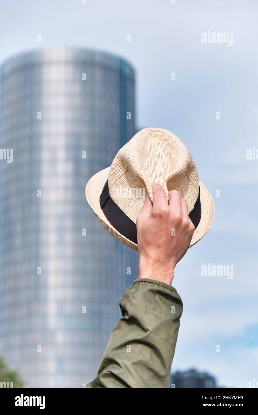 Man's hat in hand against the backdrop of a skyscraper office building and blue sky. Welcome and greeting hand gesture, goodbye office and freedom Stock Photo