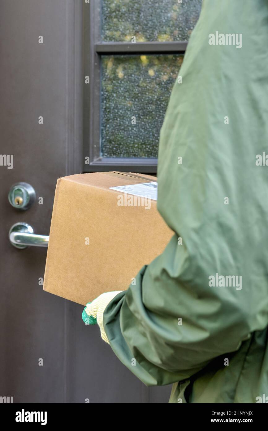 Cardboard box in the hands of the postman on the background of the door of the house. Delivery of the parcel courier service to your home. Stock Photo