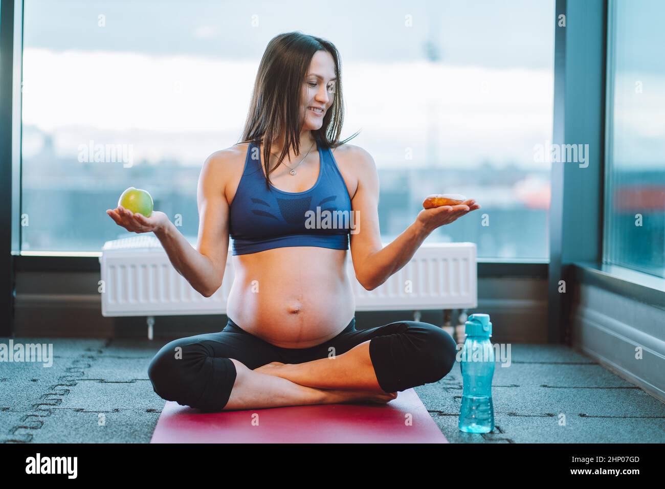 Smiling pregnant woman sportswear, yoga lotus exercise, choise apple, donut  Stock Photo