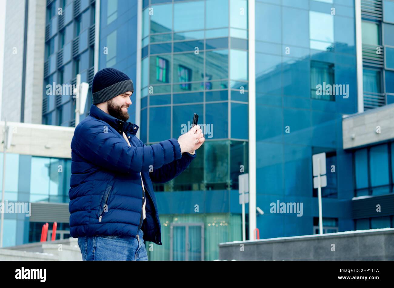 young elegant business man in winter casual clothes, jacket, hat. Walking in the city street, talking on the phone, making selfie. Working outdoor. Stock Photo