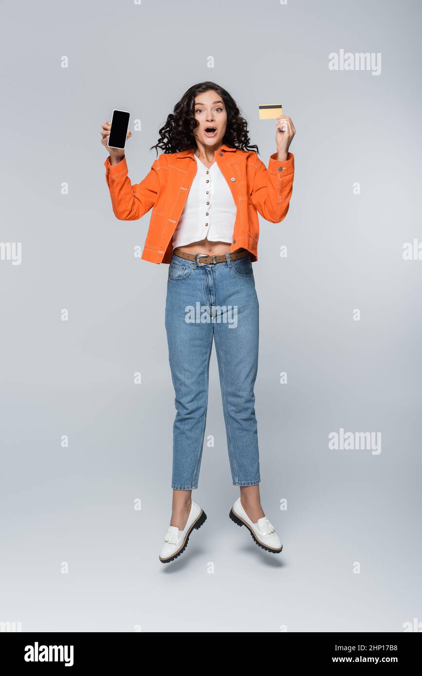 full length of shocked woman in orange jacket holding credit card with cashback and smartphone while levitating on grey Stock Photo