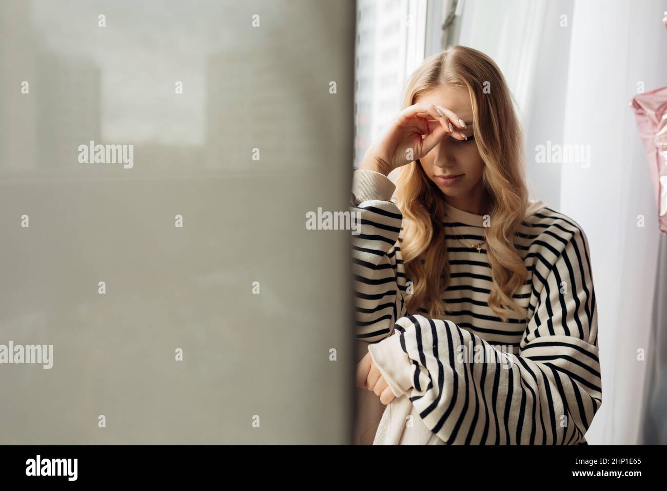 Portrait of pensive thoughtful blond pretty sad, upset, depressed woman sit on window sill, face hands. Covid lockdown Stock Photo