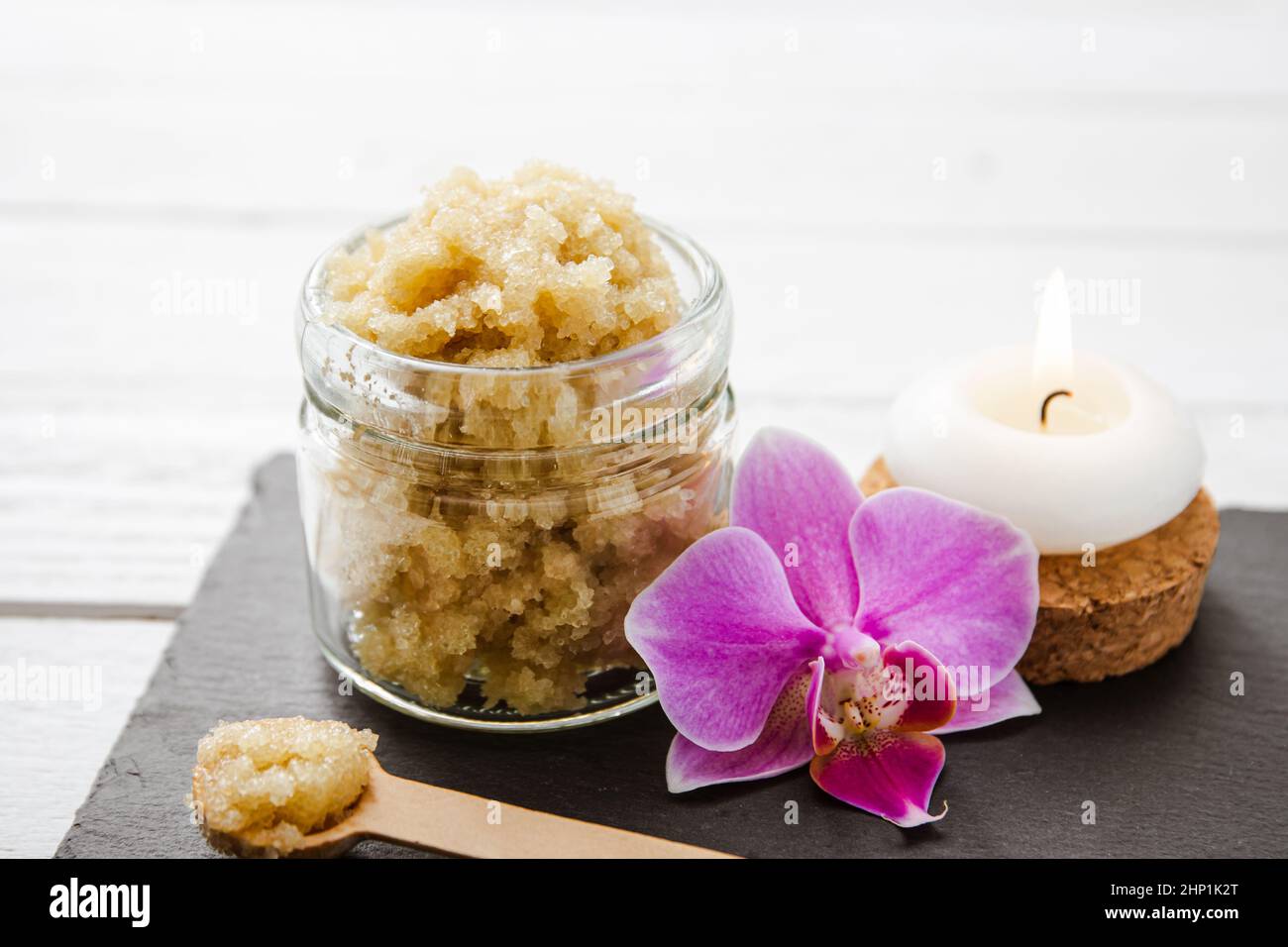 Yellow color handmade sugar body scrub in glass jar. Burning candle and pink orchid blossom for decoration on white studio background. Copy space. Stock Photo