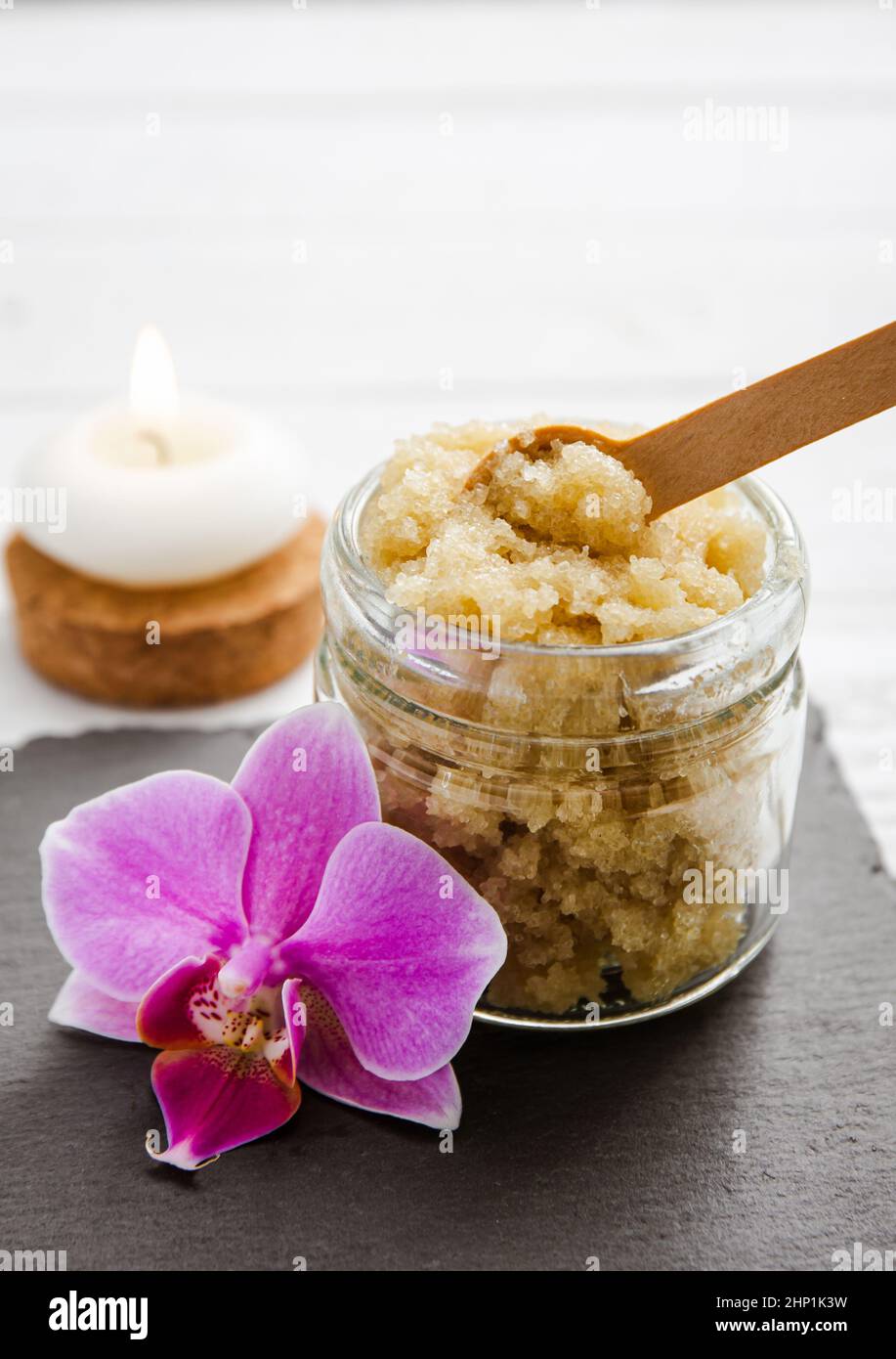 Yellow color handmade sugar body scrub in glass jar. Burning candle and pink orchid blossom for decoration on white studio background. Copy space. Stock Photo