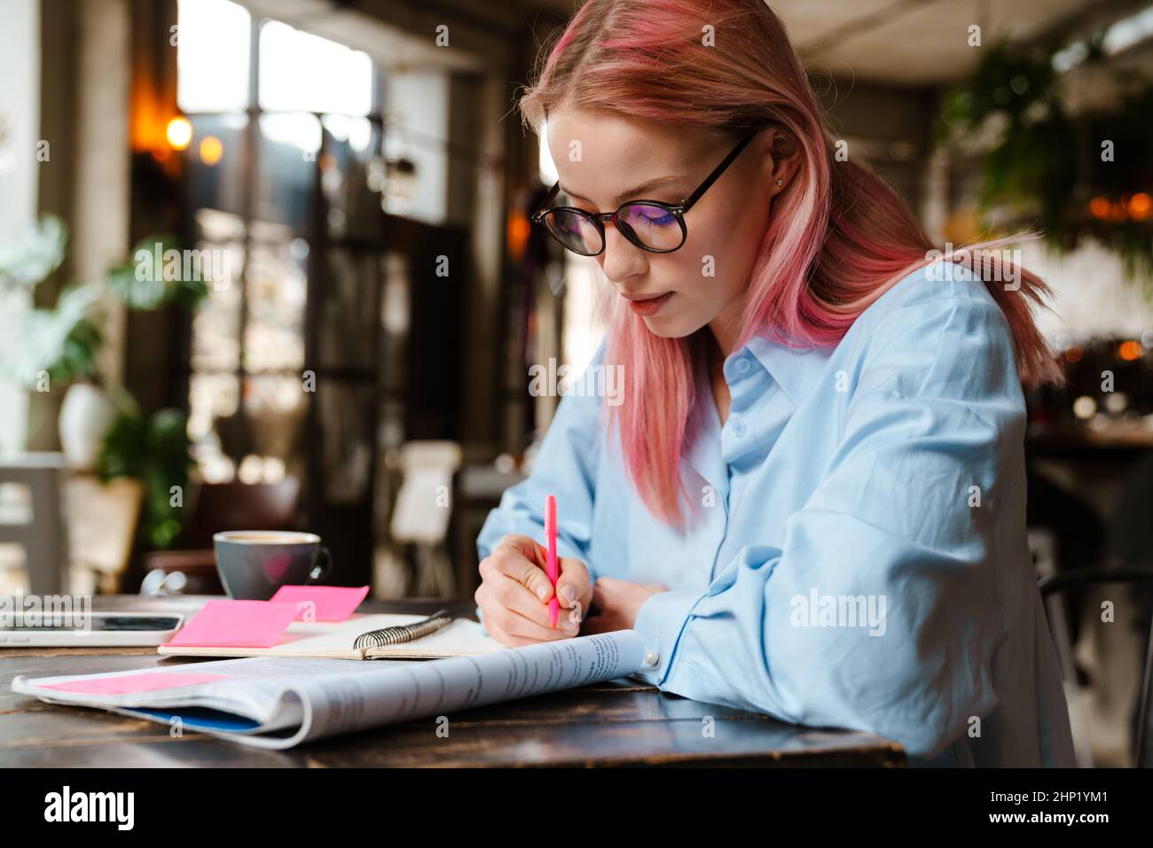 Young beautiful woman with pink hair studying and making notes while sitting in cafe Stock Photo