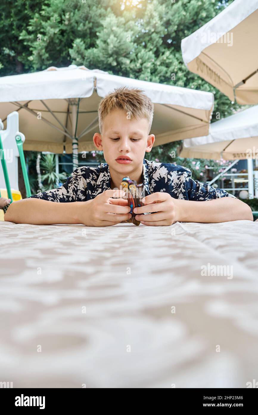 Fair-haired schoolboy in colorful shirt plays sitting at table waiting for food. Child sits in boredom near canopies against green trees closeup Stock Photo