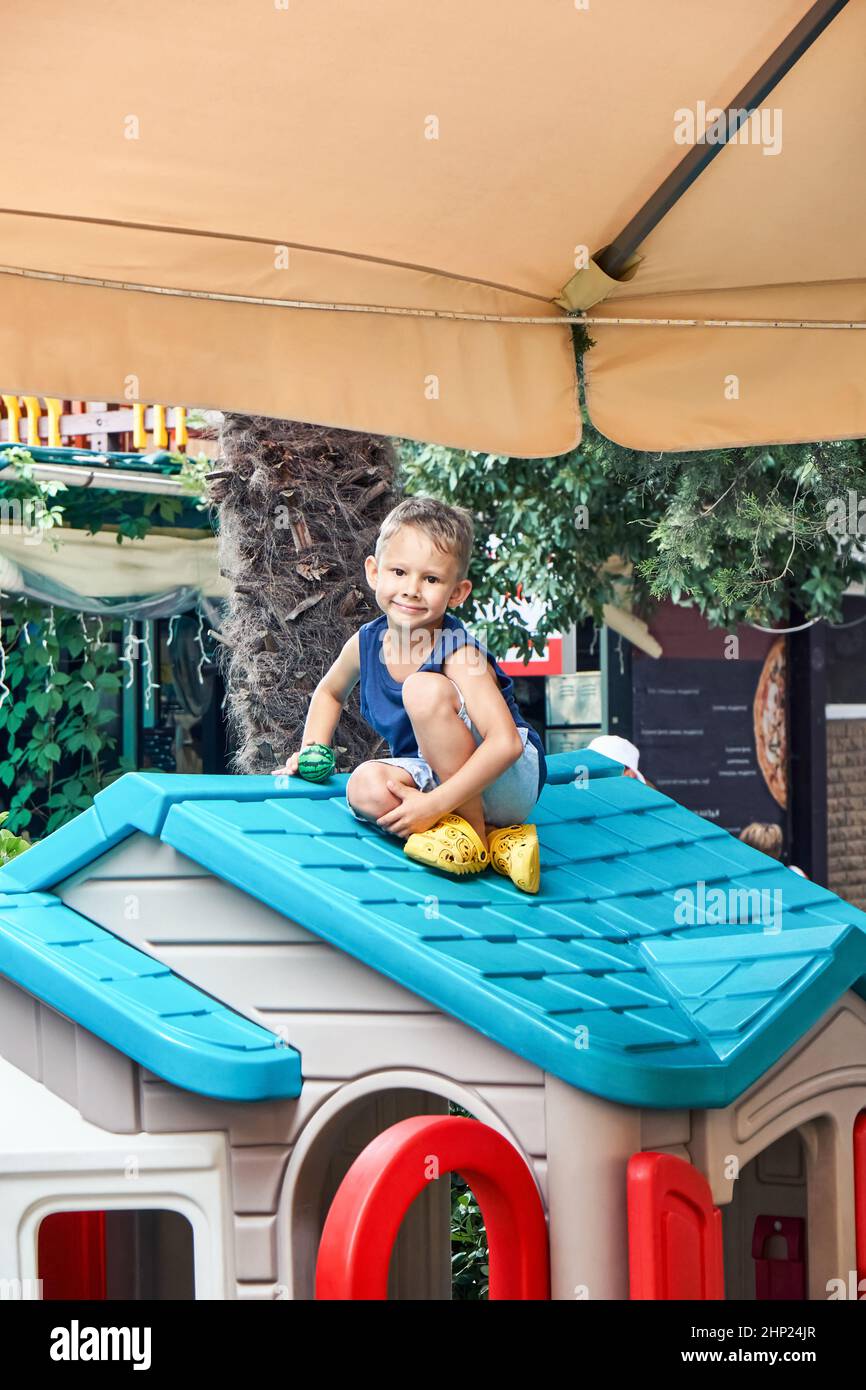 Preschooler boy in summer clothes sits on bright roof of toy house in restaurant play area. Fair-haired child spends free time playing outdoors Stock Photo