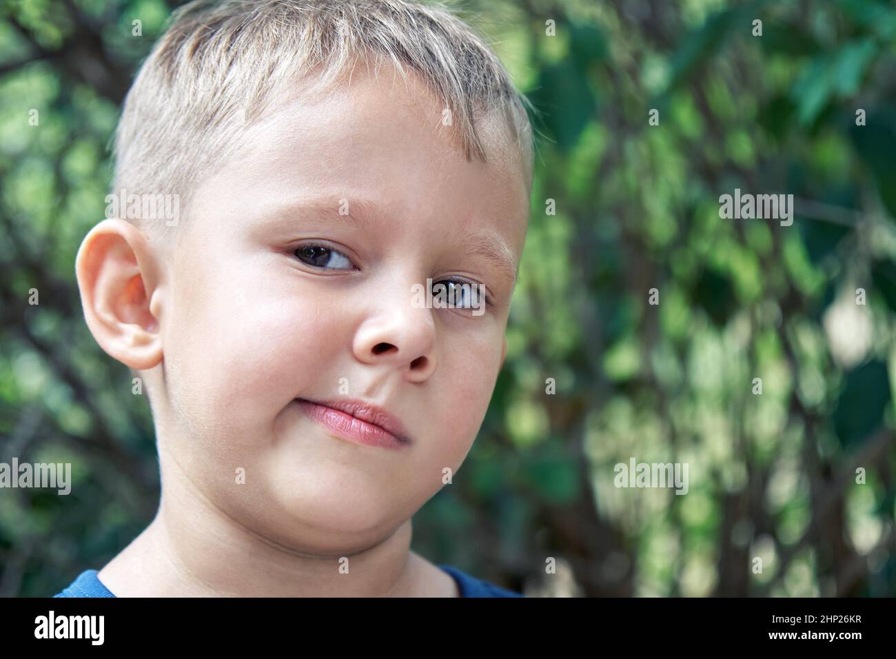 Blond preschooler grimaces making weird face expressions. Boy has fun walking outdoors in shade of trees in park on summer holiday closeup Stock Photo