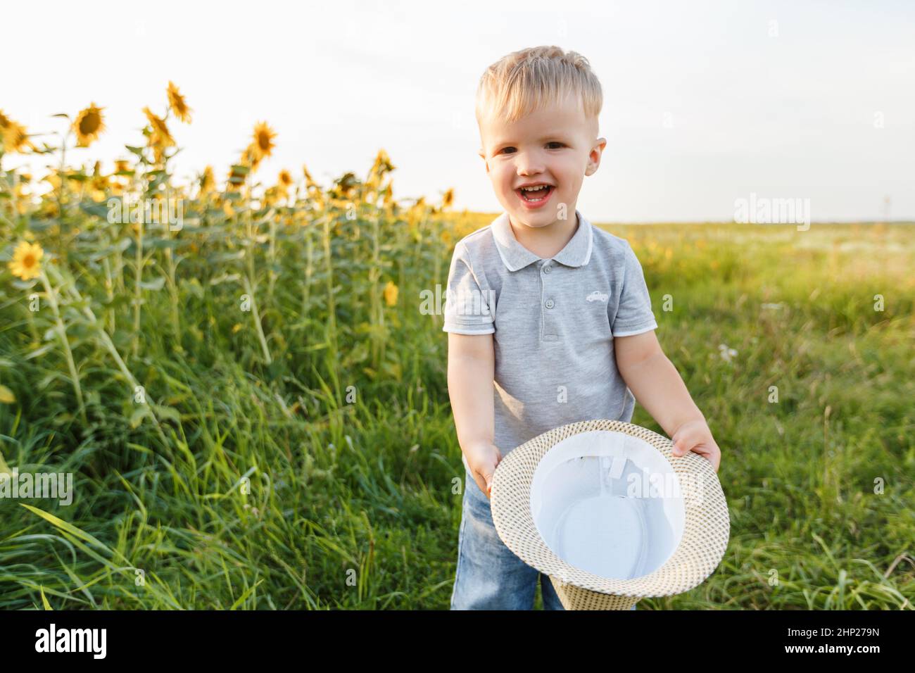 Little boy smiling, holding hat in hands and playing on the background of countryside. Three years kid having fun in field of sunflowers on sunset. Su Stock Photo