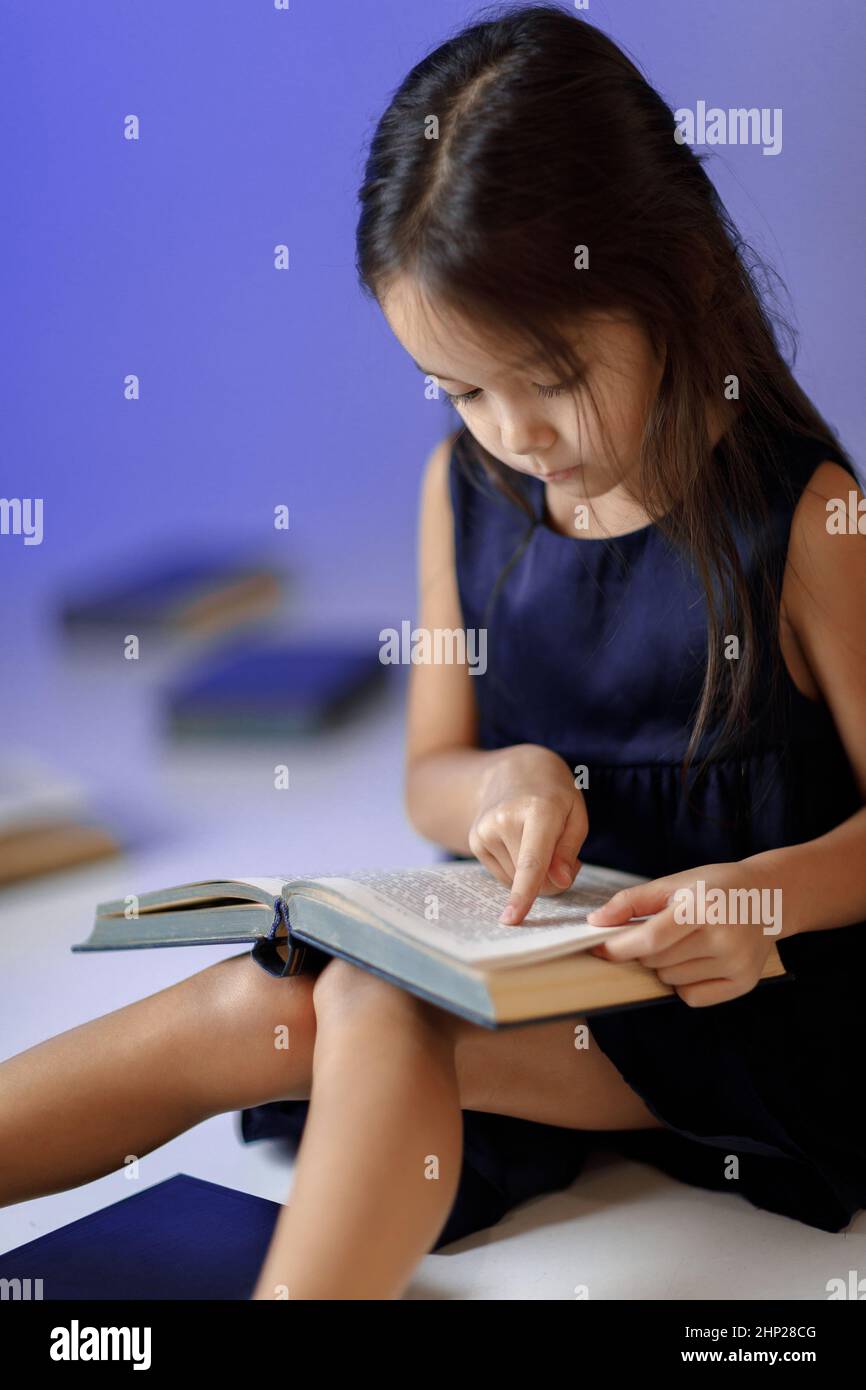 portrait of cute little girl in dark blue dress is reading a book ...