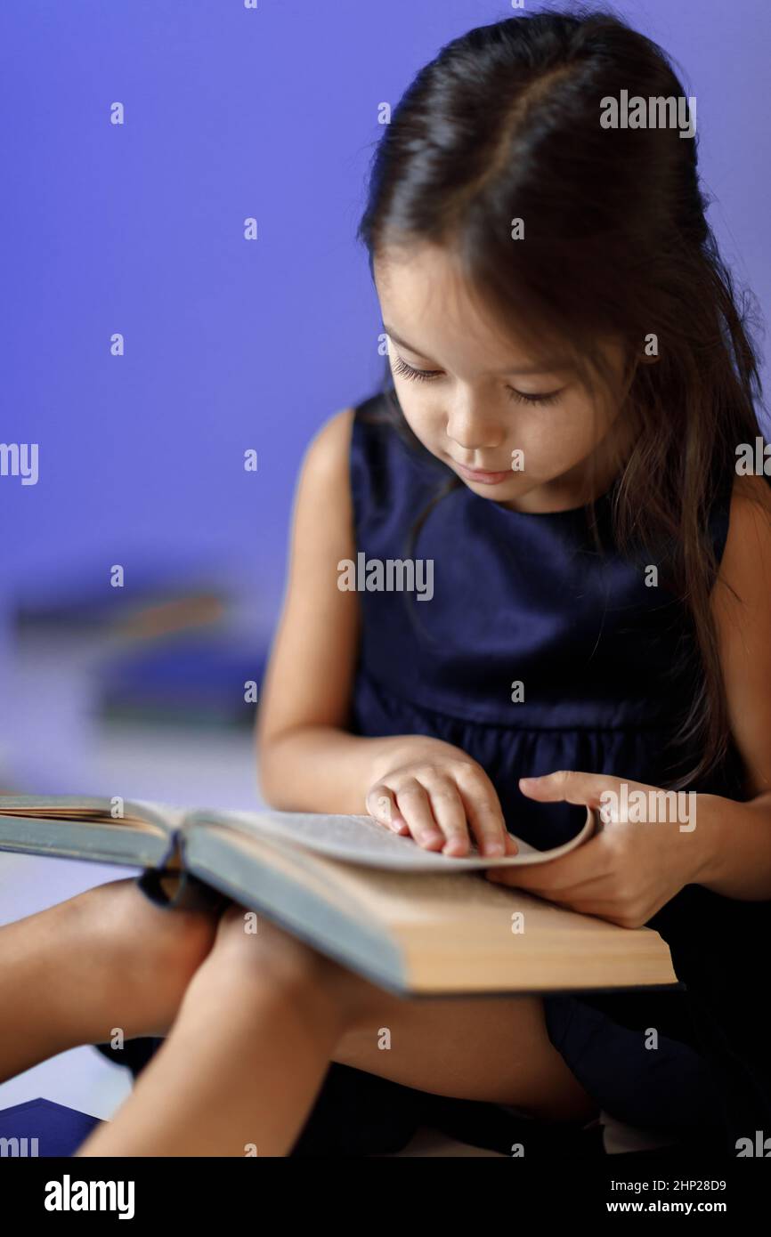 portrait of cute little girl in dark blue dress is reading a book ...