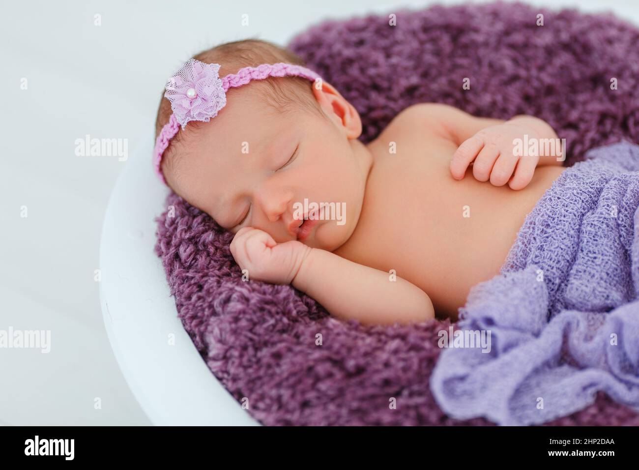 Newborn baby in violet knitted suit sleep in wooden basket. Child resting in cute pose. Tiny girl with tender headband on head. Creative infant photog Stock Photo