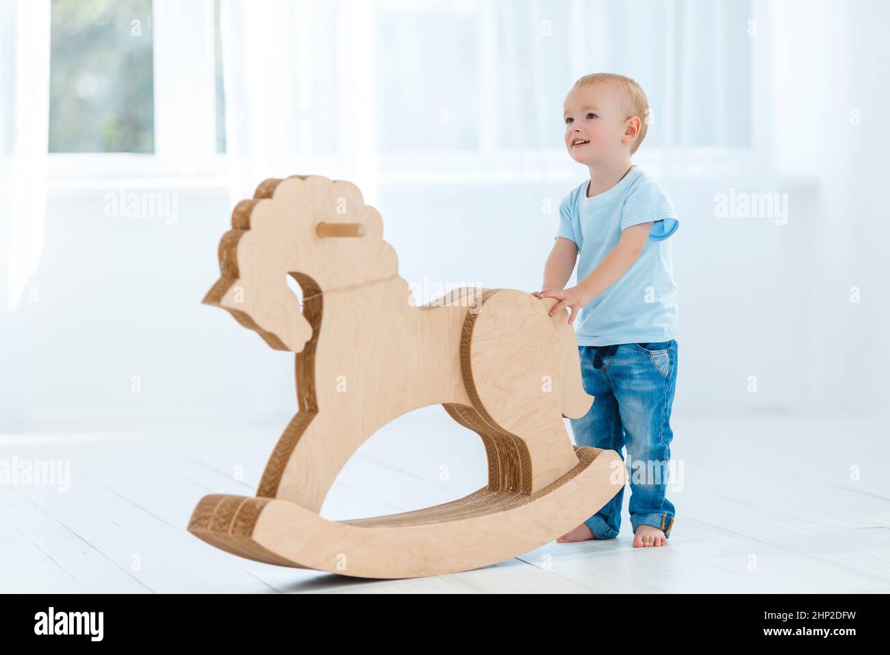 Cute, smiling, white, two years old boy in blue t-shirt and jeans rocking on wooden handmade horse. Little child having fun with pony toy. Concept of Stock Photo