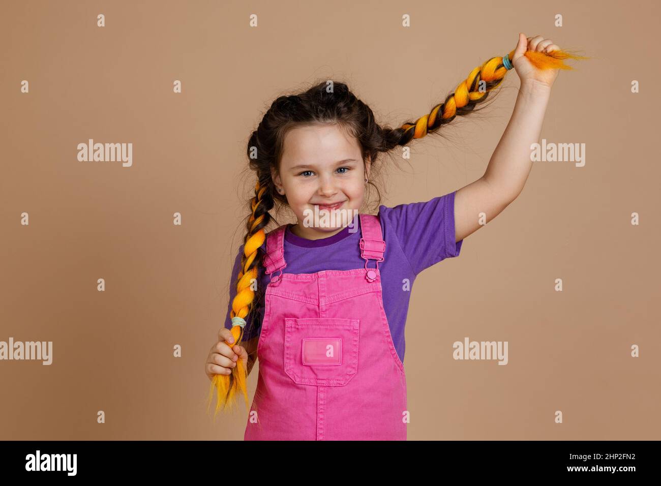 Beautiful, little girl, pulling kanekalon pigtails of yellow color, looking at camera with gentle smile, wearing pink jumpsuit and purple t-shirt on Stock Photo