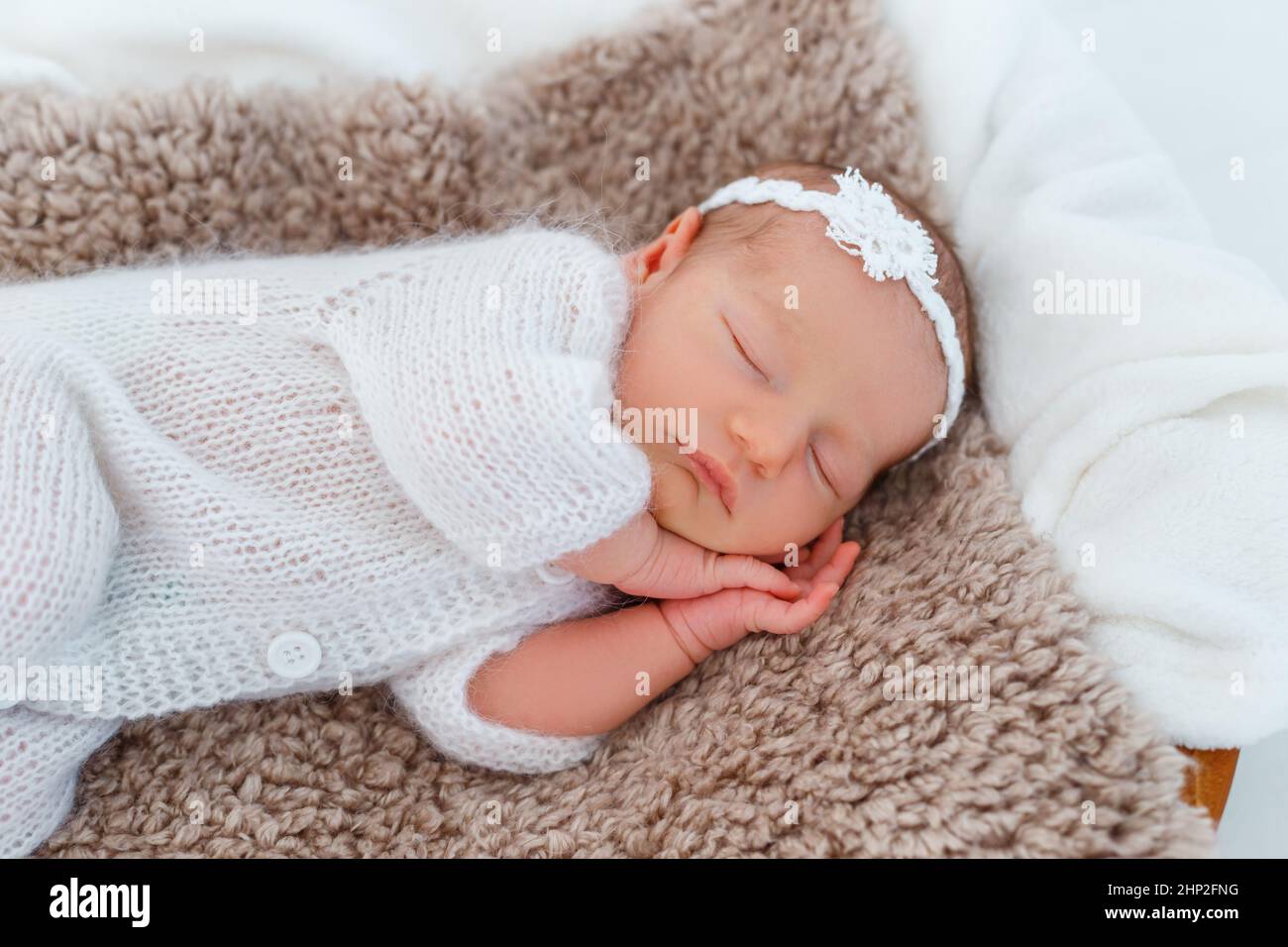 Newborn baby in white knitted suit sleep in wooden basket. Child resting in cute pose. Tiny girl with tender headband on head. Creative infant photogr Stock Photo