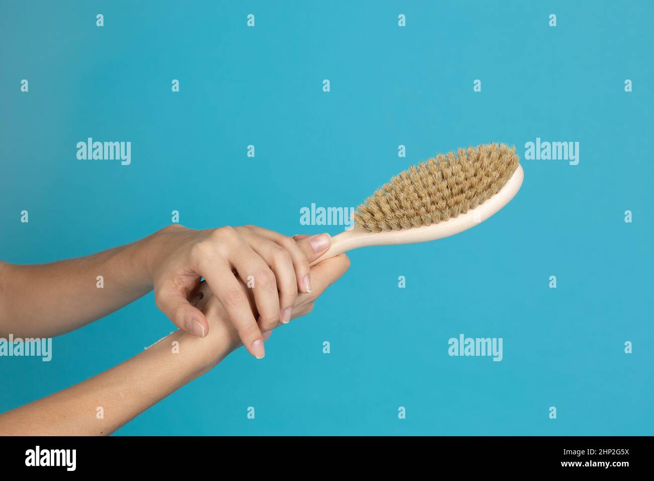 Two hands with wooden brush for massage of dry skin on blue background. Anti-cellulite procedures aiming for getting rid of cellulite. Self-care and Stock Photo