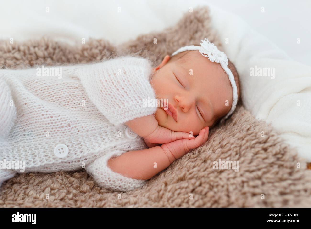 Newborn baby in white knitted suit sleep in wooden basket. Child resting in cute pose. Tiny girl with tender headband on head. Creative infant photogr Stock Photo