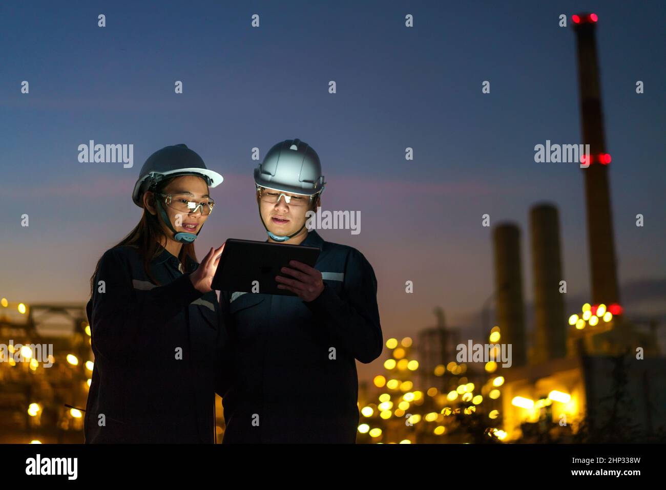 Asian engineers, man and woman are checking the maintenance of the oil refinery factory at night via digital tablets. Stock Photo