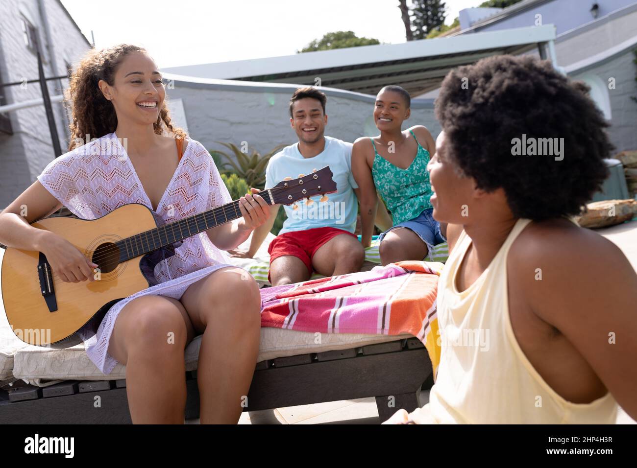 Smiling young multiracial woman playing guitar in front of friends sitting at poolside Stock Photo