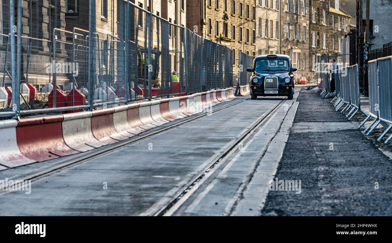 Black taxi cab driving on tram tracks with roadworks barrier, Constitution Street, Leith, Edinburgh, Scotland, UK Stock Photo