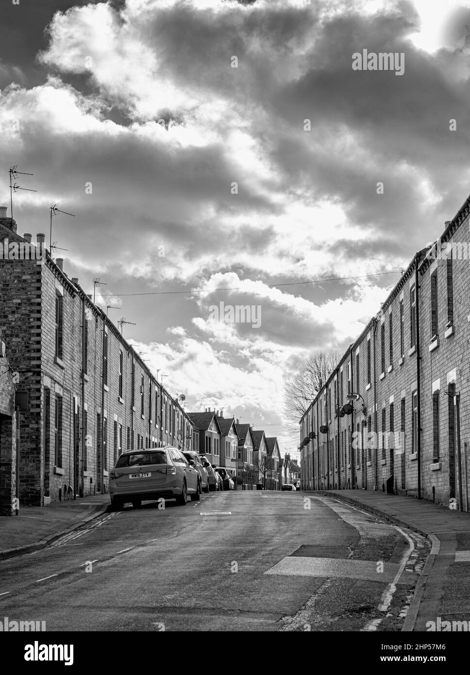 A street with neat terraced houses are situated on a rise, sloping upwards. Cars are parked on one side and a cloudy sky is above Stock Photo