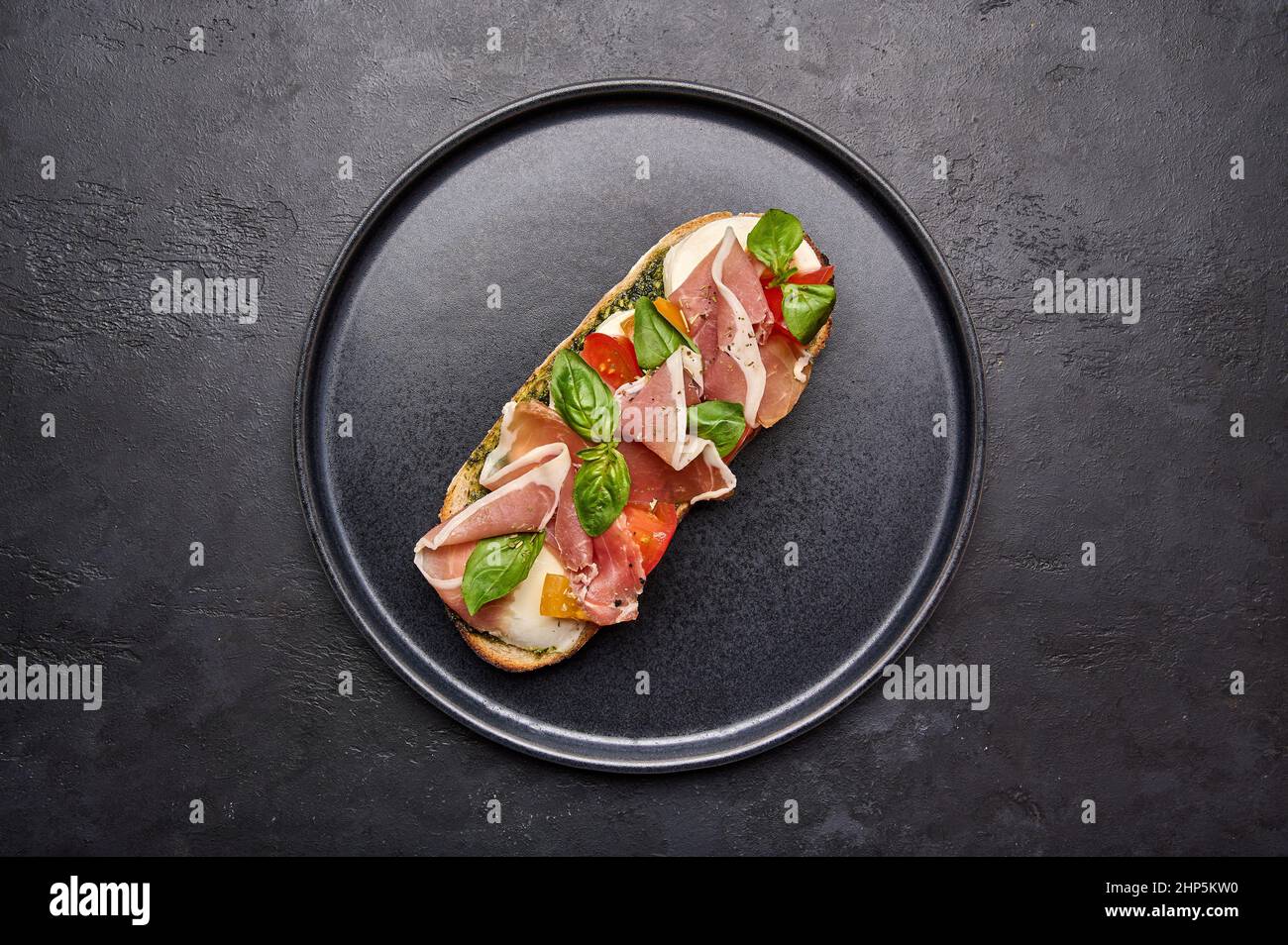 Top view bruschetta with mozzarella, Parma ham and pesto on black plate on graphite background Stock Photo