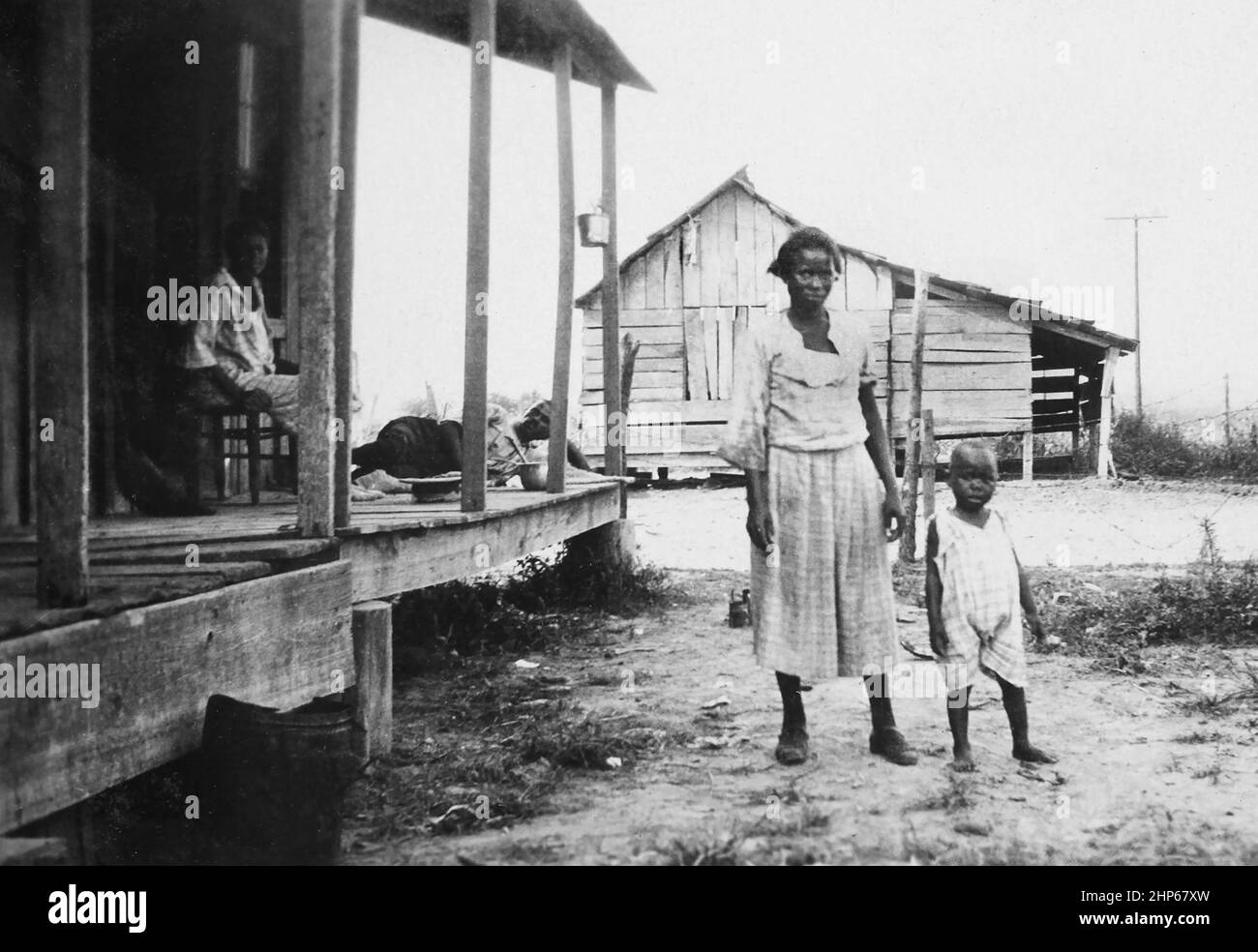 A young mother stands outside her sharecropper shack with her two children while the father sleep on the porch in Mississippi.,  ca. 1925. Stock Photo