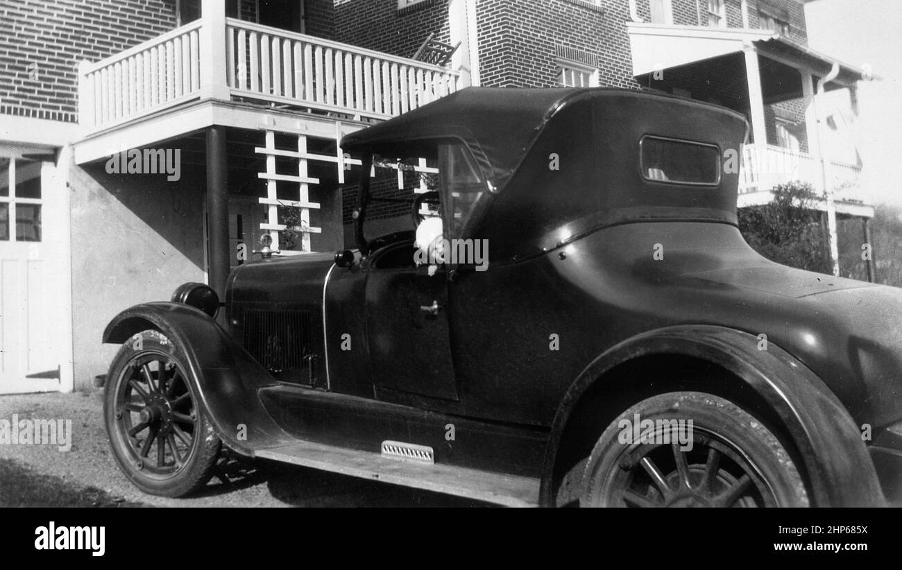 A little girl peeks out a window of stylish 1920s coupe automobile in the driveway, ca. 1928. Stock Photo