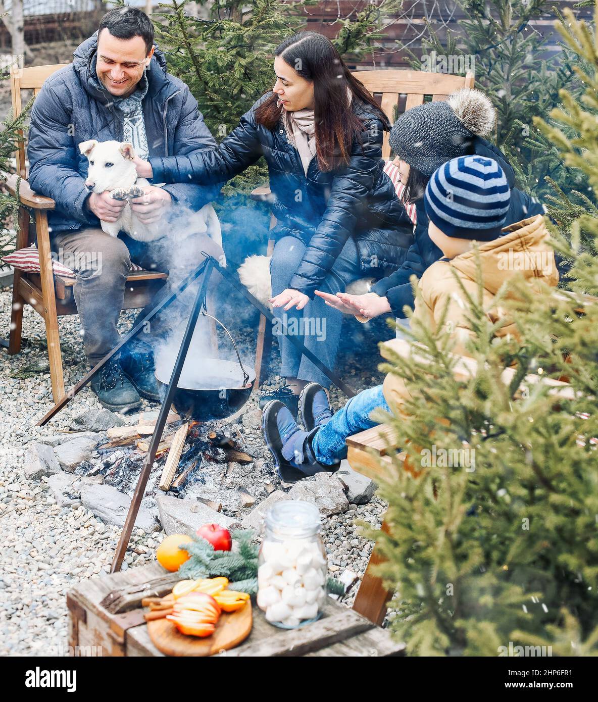 New Years gatherings by winter bonfire. Cozy portrait of happy family parents, two children and sweet dog cooking food in steaming pot over campfire w Stock Photo