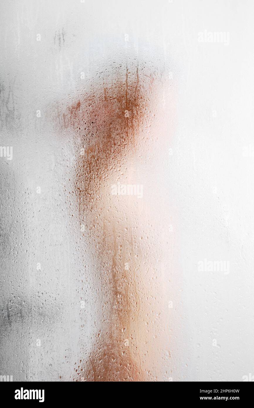A woman with blond hair takes a shower through fogged glass with water drops neutral background Stock Photo