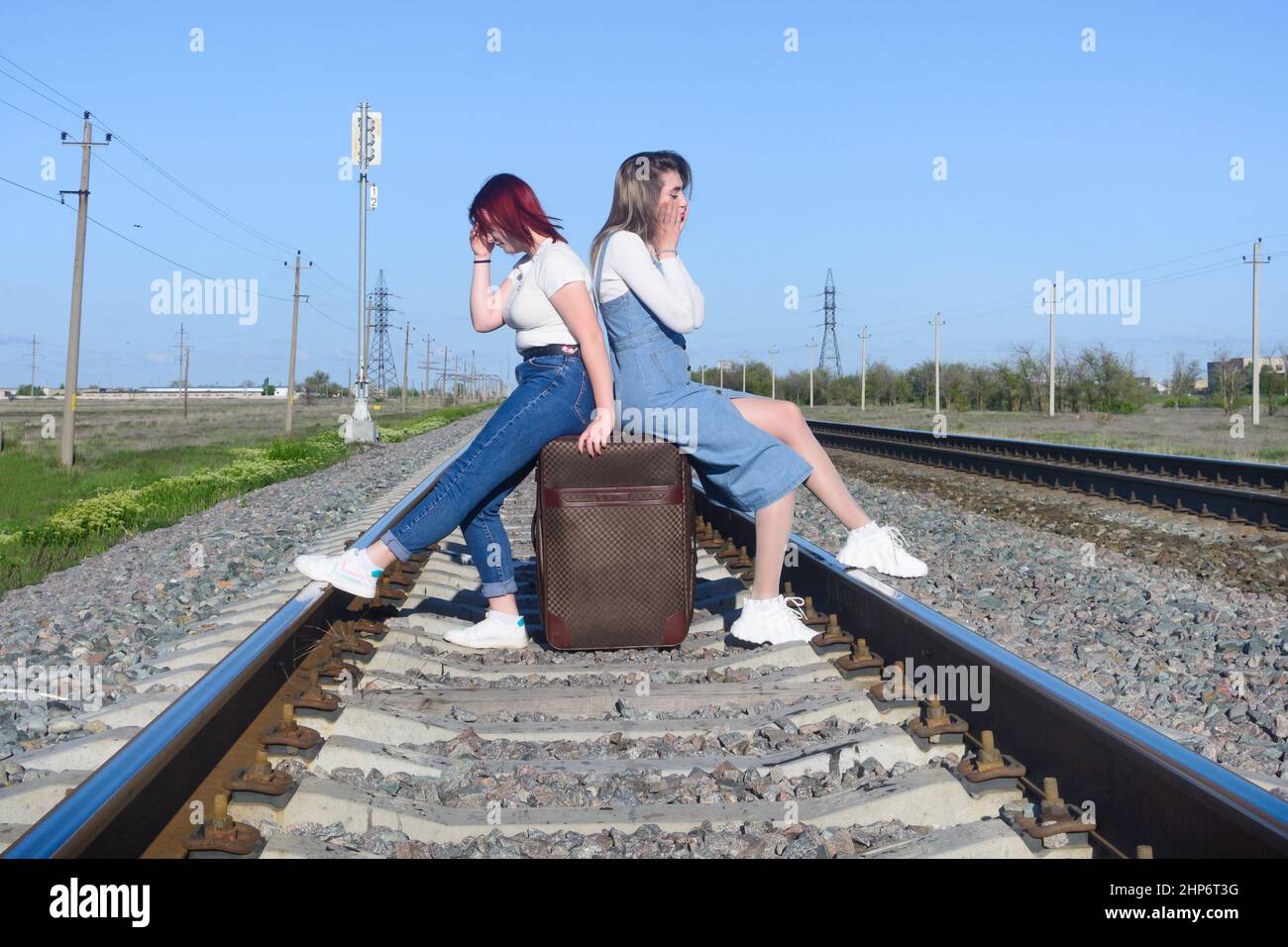 Beautiful tired young ladies sit on large suitcase on the railroad tracks. Difficult travel concept. Stock Photo