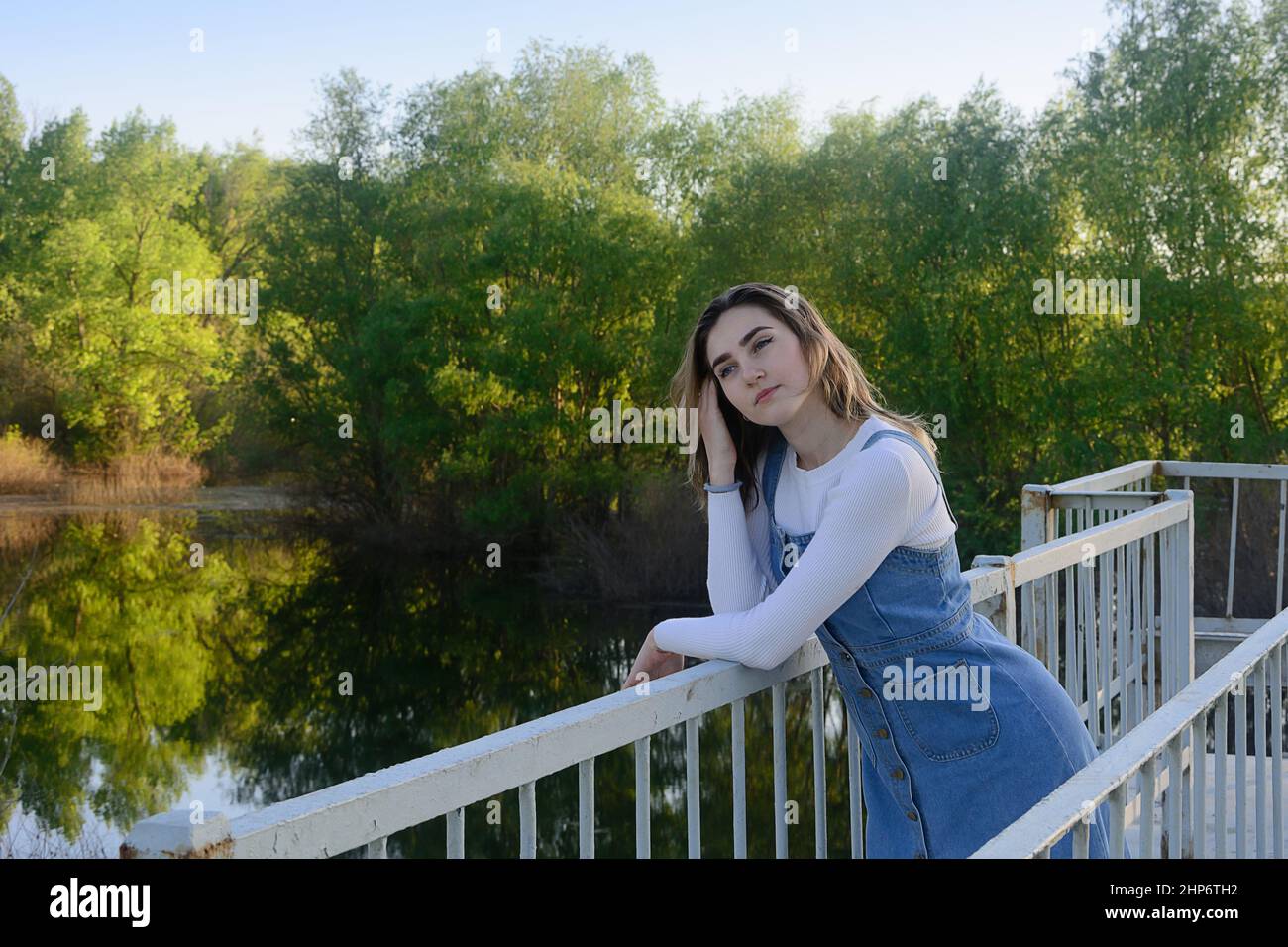 Beautiful young woman stands near railing on the bridge over river. Romantic summer mood. Stock Photo