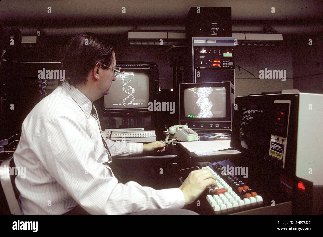 A Caucasian male technician working at a computer terminal and consulting several other screens. This technology allows for the storage and retrieval of vast amounts of information needed for cancer research. DNA structures are on the screen ca.  1980 Stock Photo