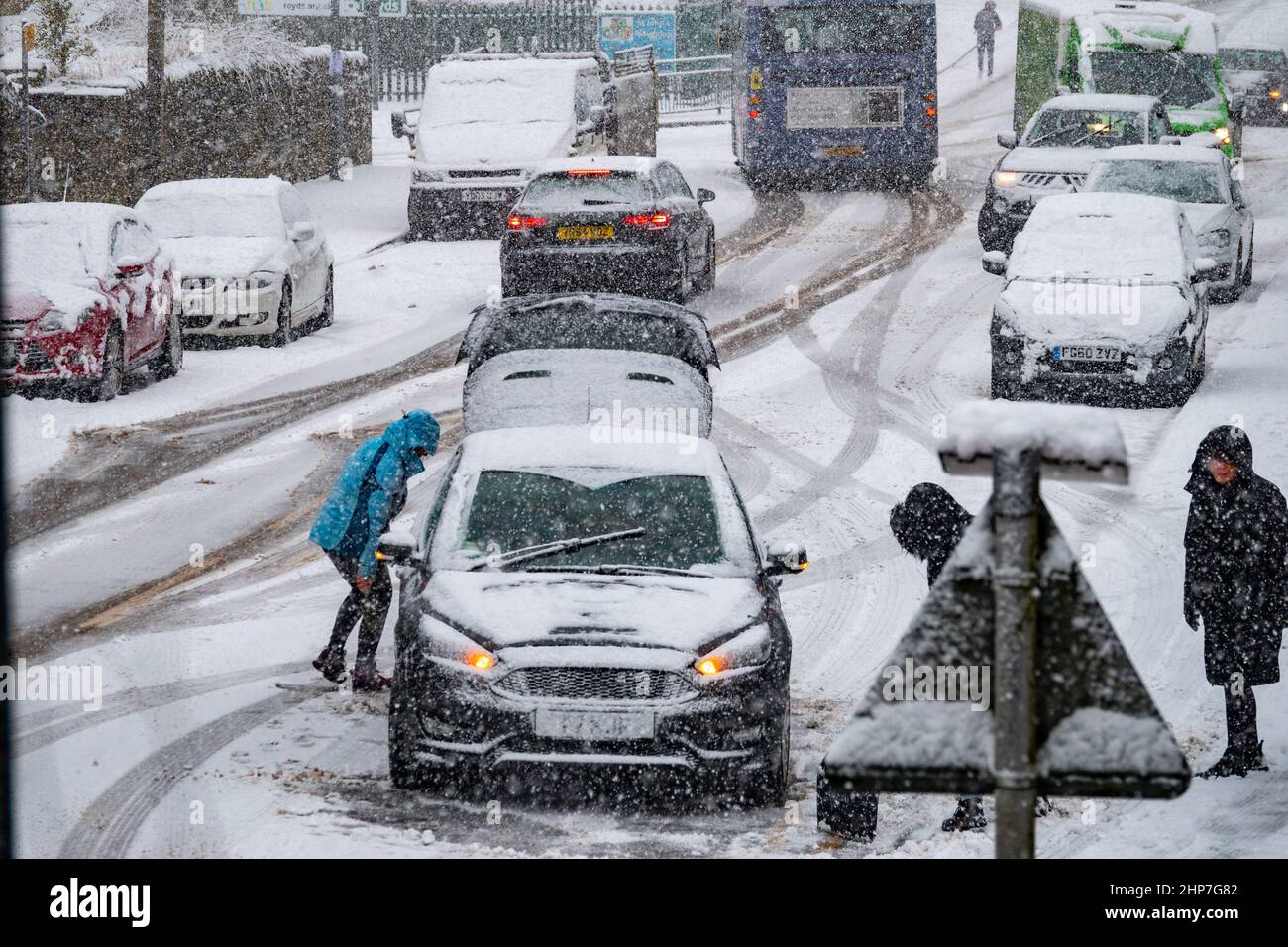 Snow and ice causing disruption on the hills around Bradford, West Yorkshire, UK. 19th Feb 2022. Cars struggle to move on slippery winter surfaces. Women try to clear wheels on snowy hill to get their car moving. Stock Photo