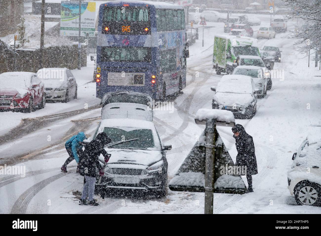 Snow and ice causing disruption on the hills around Bradford, West Yorkshire, UK. 19th Feb 2022. Cars struggle to move on slippery winter surfaces. Women try to clear wheels on snowy hill to get their car moving. Stock Photo