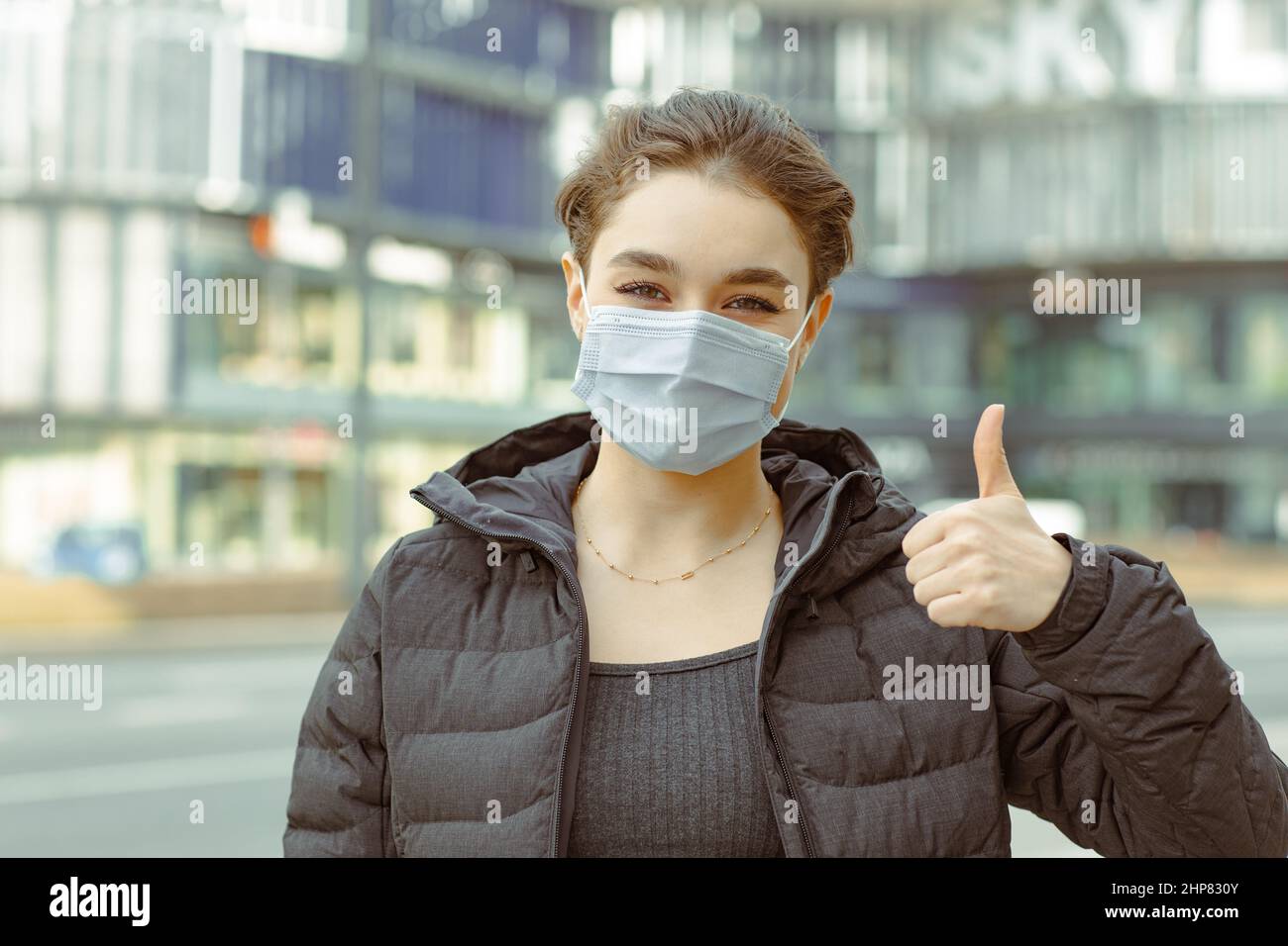 Portrait of a young woman wearing a face mask standing on a street giving a thumbs up gesture Stock Photo