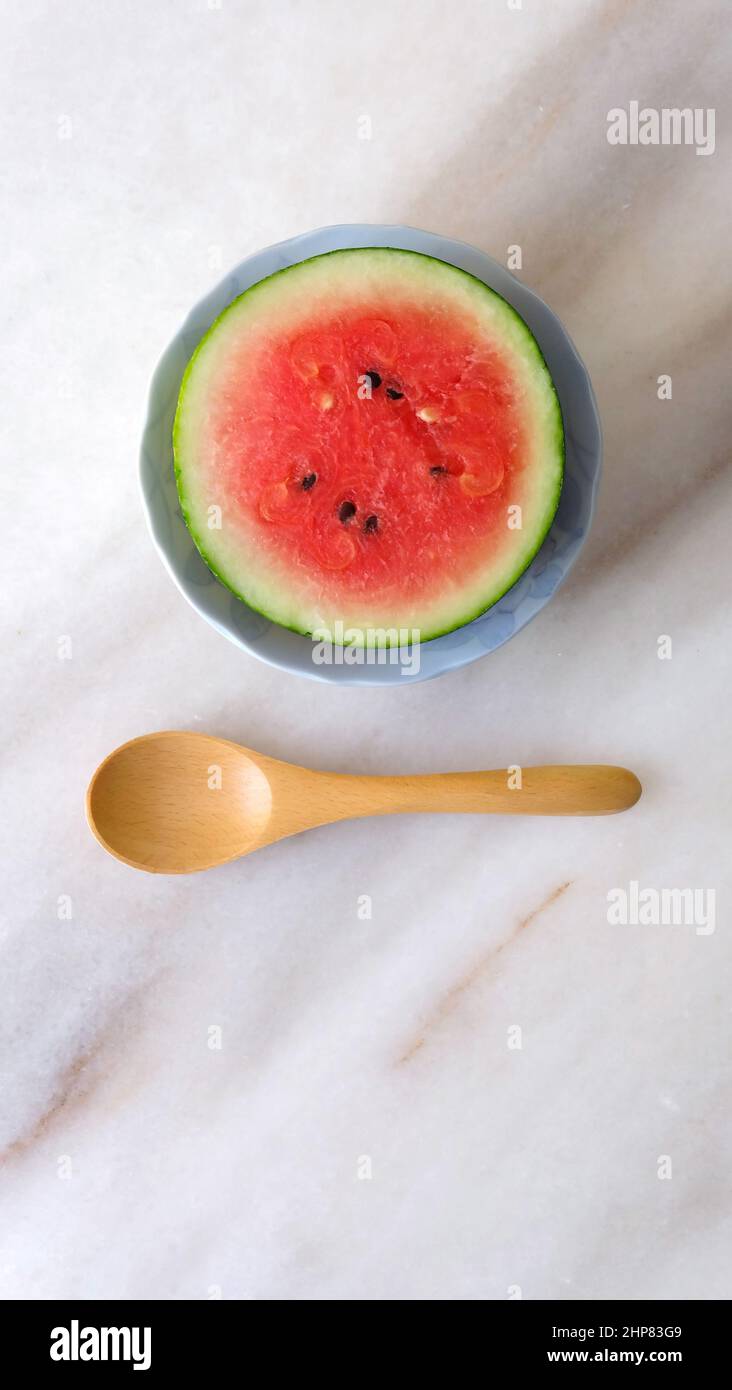 Flat lay of a small round watermelon cut in half, and a wooden spoon next to it. Placed on a marble counter top. Stock Photo