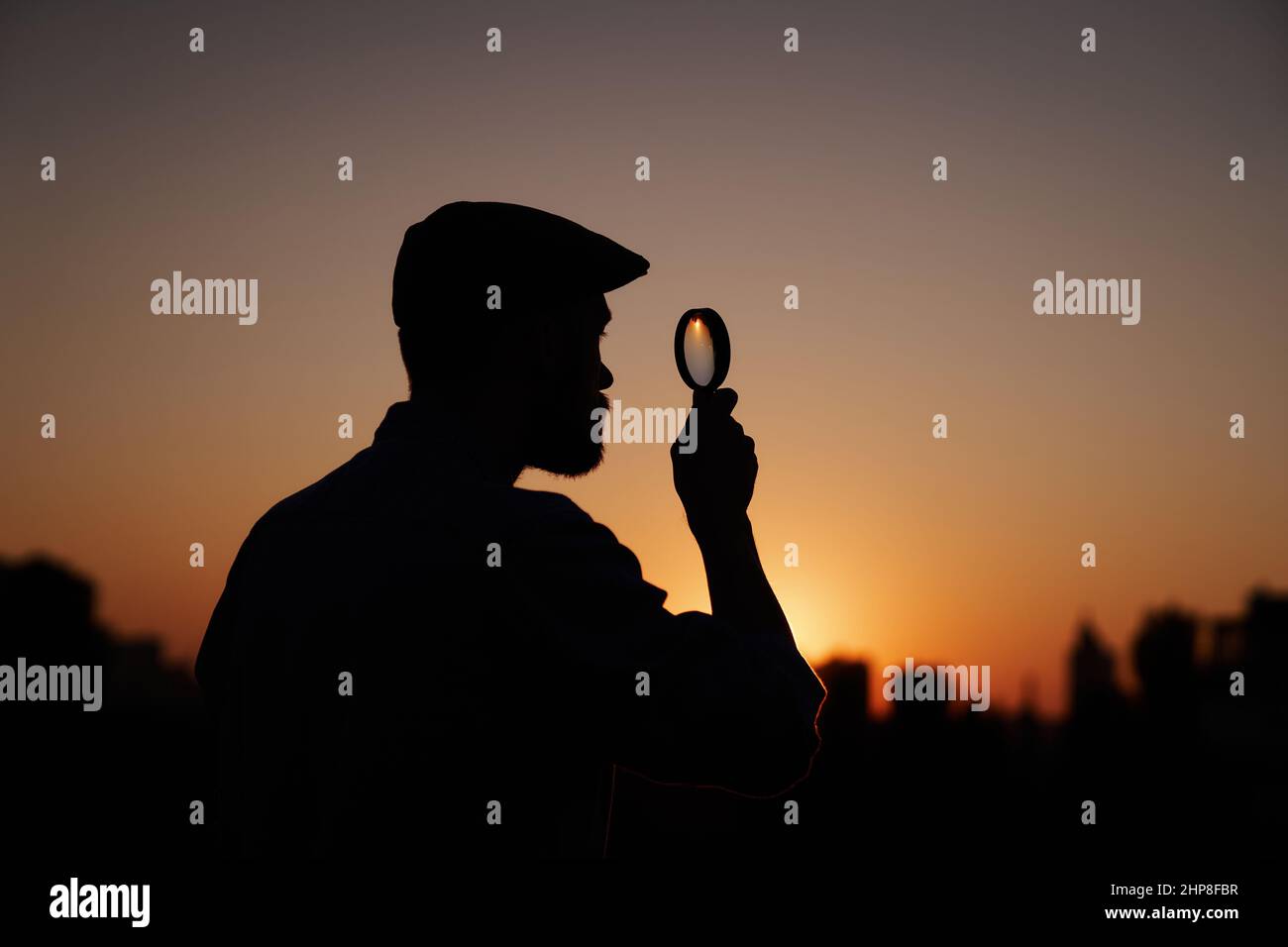 Male detective or investigator silhouette portrait in peaked hat looking with magnifying at side. Man searching using loupe with sun on background and urban view. Investigation or inspection concept Stock Photo