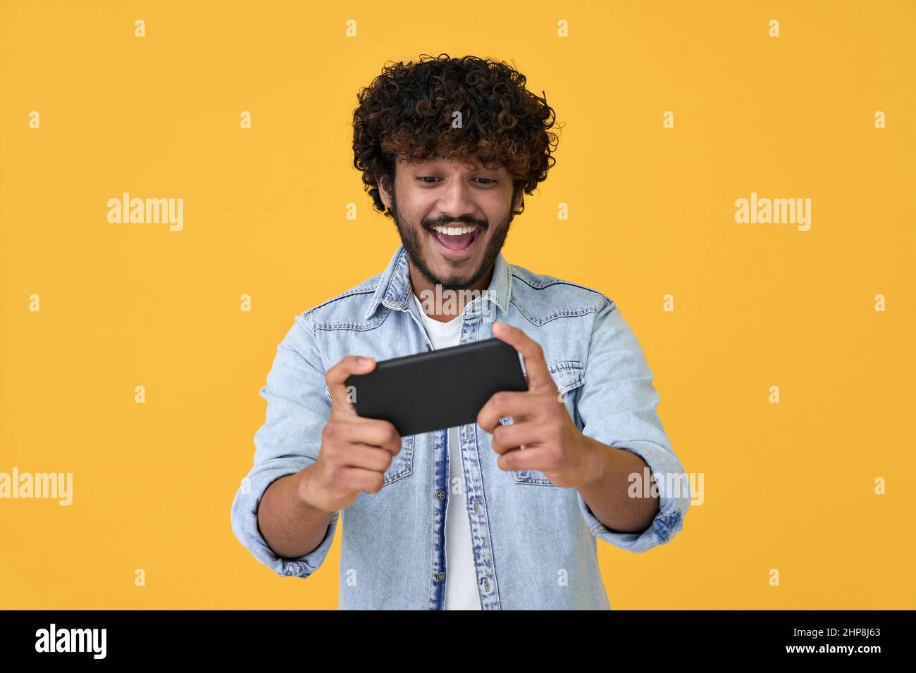 Excited young indian man using smartphone playing game on yellow background. Stock Photo