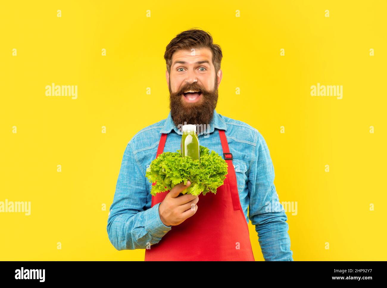 Surprised man in apron holding fresh lettuce and juice bottle yellow background, juice barkeeper Stock Photo