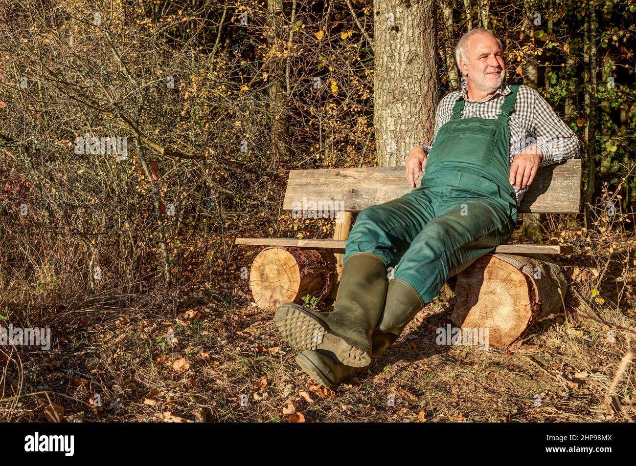 A farmer with green dungarees and rubber boots relaxes after work on his bench at the edge of the forest in the mild evening sun. Stock Photo