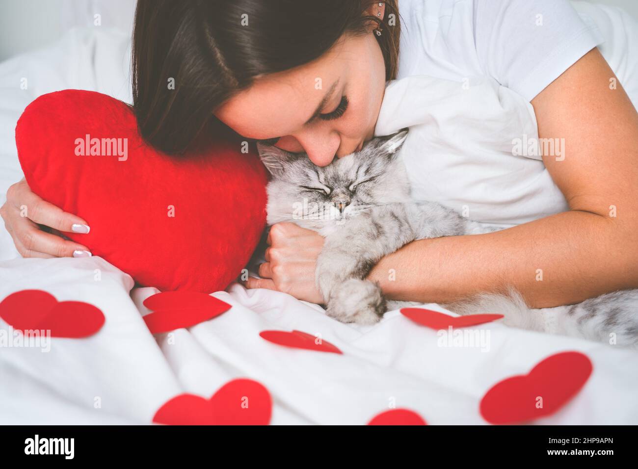 Woman lying on the bed with red hearts and kissing her lovely cat. Love for pets. Valentine's day concept Stock Photo