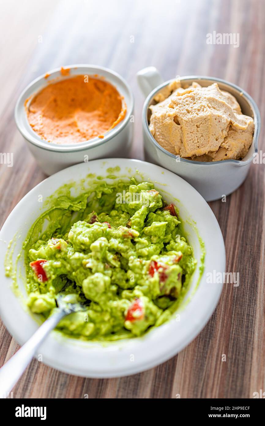 Above closeup view of guacamole dish in white bowl made with ripe green avocado, tomatoes and chickpea hummus red pepper spread as snack lunch food se Stock Photo