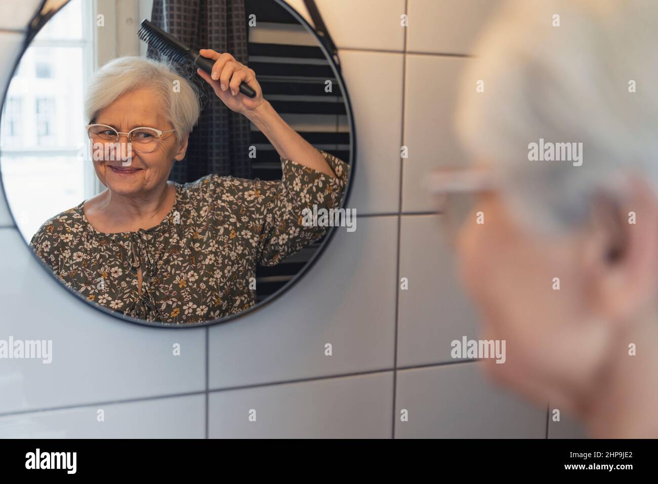 senior grey-haired woman brushing her hair and looking at her reflection in the mirror . High quality photo Stock Photo